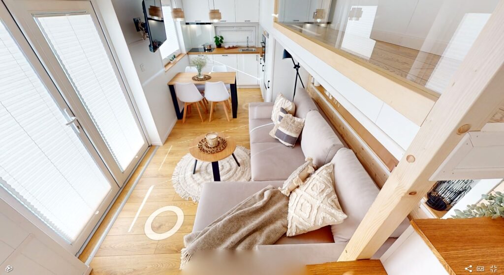Capture d'écran d'une visite virtuelle interactive de la tiny house, offrant une vue panoramique à 360 degrés de l'intérieur moderne et écologique. L'image montre une interface utilisateur intuitive avec des flèches de navigation et des points d'information, permettant aux visiteurs de cliquer et d'explorer différentes parties de la maison, comme la cuisine, la mezzanine, et l'espace de vie. La technologie de réalité virtuelle permet une immersion complète, donnant l'impression de se déplacer réellement à l'intérieur de cette maison compacte mais élégamment conçue.